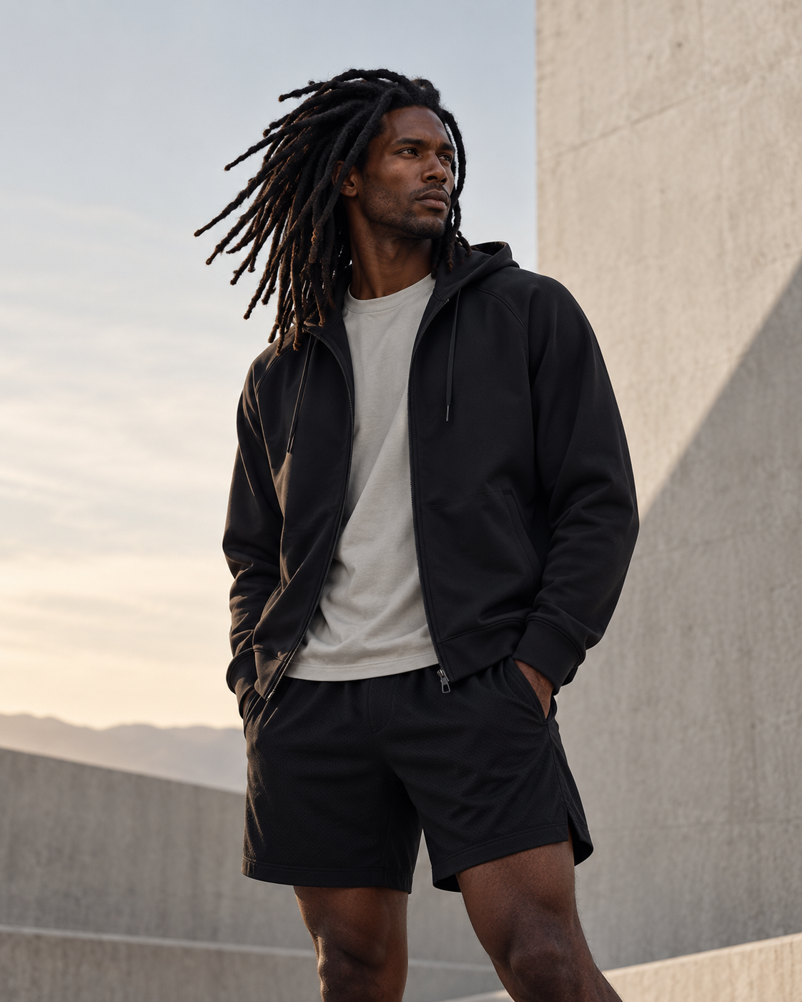 Model wearing black zip hoodie and black athletic shorts against a concrete architectural backdrop